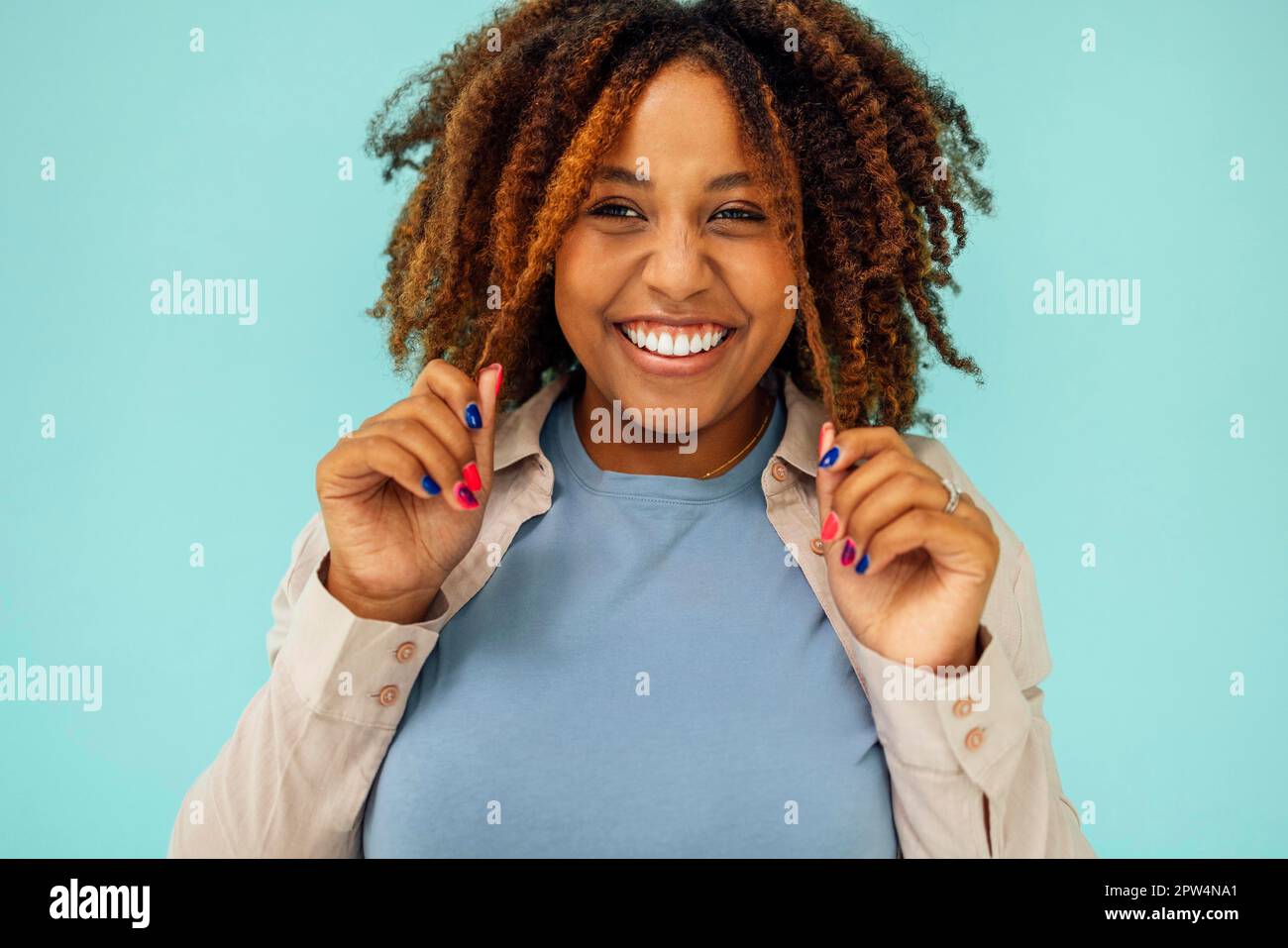Positive afroamerikanische weibliche Model lächelt an der Kamera und berührt sauberes lockiges Haar nach der Hygienebehandlung auf blauem Hintergrund Stockfoto