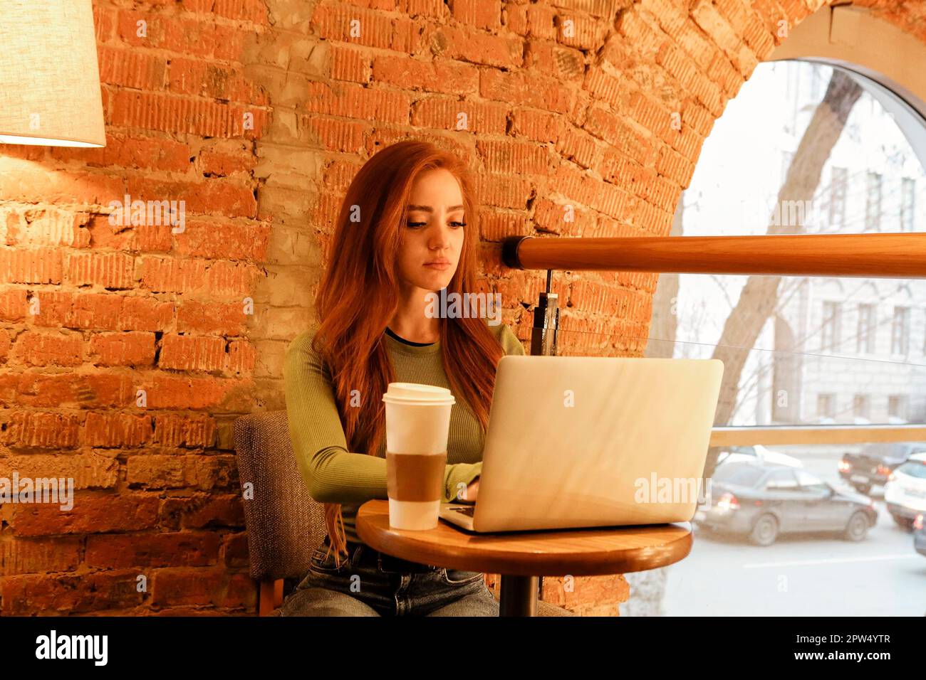 Junge Rothaarige Frau mit Kaffee, um am Tisch sitzen zu gehen In der Nähe einer Ziegelwand und beim Surfen im Laptop, während Sie bei Freelance arbeiten Projekt in der Cafeteria Stockfoto