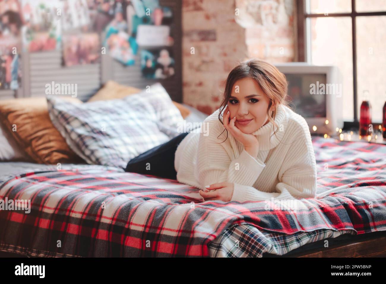 Junge Frau in einem warmen Strickpullover, die auf einem bequemen Bett mit kariertem Kariert an der Wand mit Vintage-Postern zu Hause auf die Kamera schaut Stockfoto