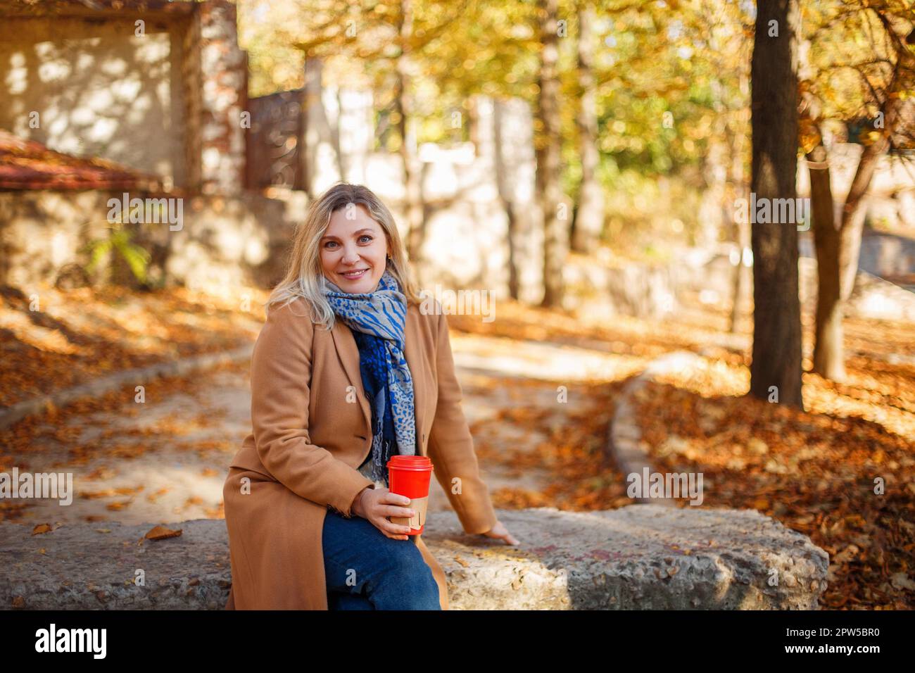 Eine wunderschöne, lächelnde blonde Frau mittleren Alters mit beigefarbenem Mantel und Schal, die am sonnigen Herbsttag im Stadtpark sitzt und ihre Tasse Kaffee zum Mitnehmen genießt. Stockfoto