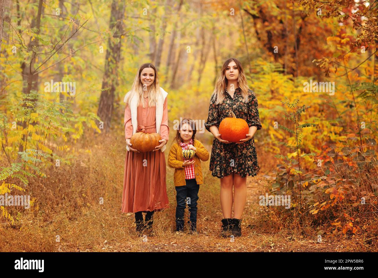 Fotos von Freunden, die draußen stehen, mit einem furchterregenden Kürbis in den Händen auf dem Weg voller heruntergefallener Blätter im Herbstpark, zwei Erwachsene Frauen und ein kleines Mädchen Stockfoto