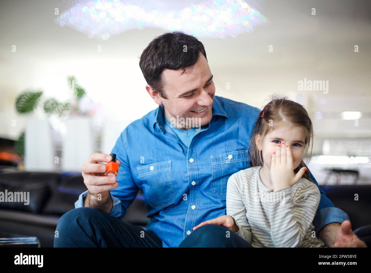 Fröhlicher Vater spielt mit der kleinen Tochter, die auf der Couch sitzt und dabei hilft, ihre Nägel mit helfender Politur zu malen. Junger brünetter Vater Stockfoto