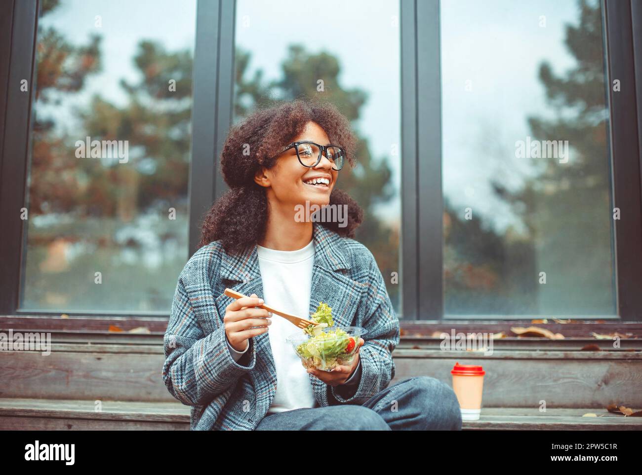 Glückliche afroamerikanische Büroangestellte, die Salat isst und vor der Kamera lächelt, während sie draußen auf einer Bank im Park sitzt, mit selektivem Fokus. Absolut Stockfoto