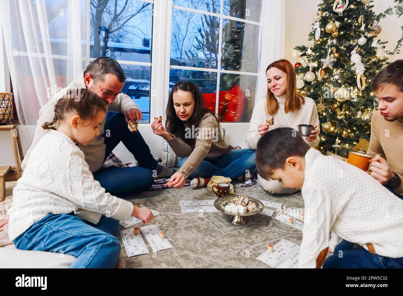 Große Familie mit Kindern, die auf dem Boden neben dem Weihnachtsbaum sitzen und gemeinsam Lotto-Brettspiele spielen, während sie an Weihnachten in gemütlichen Wohnungen verbringen Stockfoto