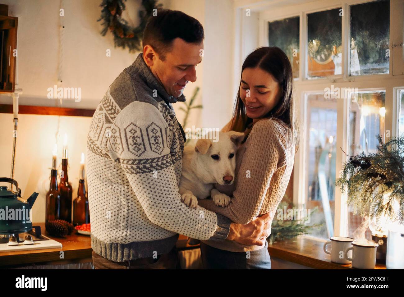 Ein junges glückliches Paar, Mann und Frau, halten einen süßen weißen Hund in der Hand und umarmen sich, während sie in der Küche stehen, dekoriert für Weihnachten, nette Familie mit Hündchen Stockfoto