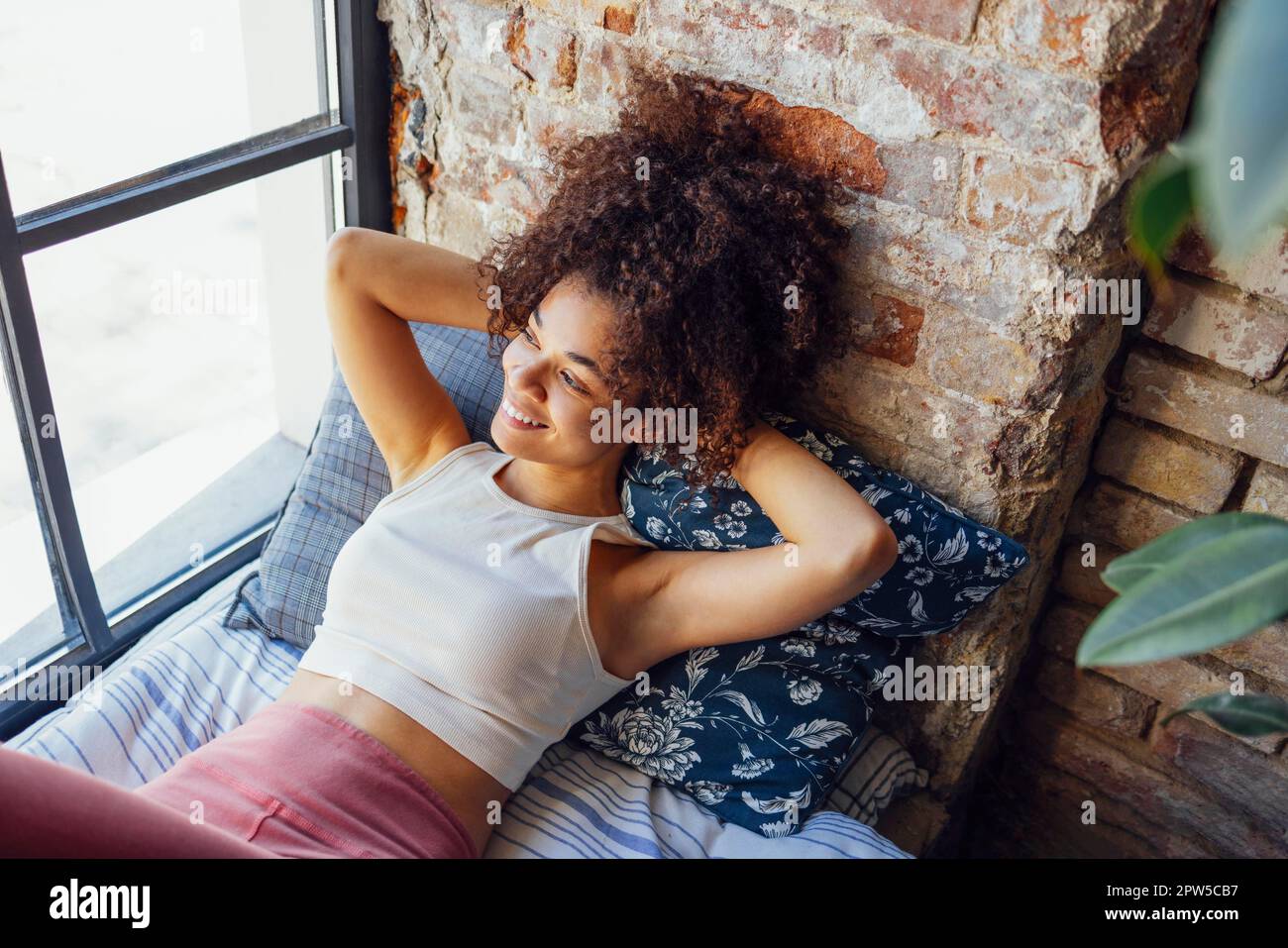 Ein entspanntes, afroamerikanisches Mädchen in Hausbekleidung, das auf einer Fensterbank sitzt, eine glückliche Afro-Frau zu Hause, die sanft zum Fenster lächelt Stockfoto