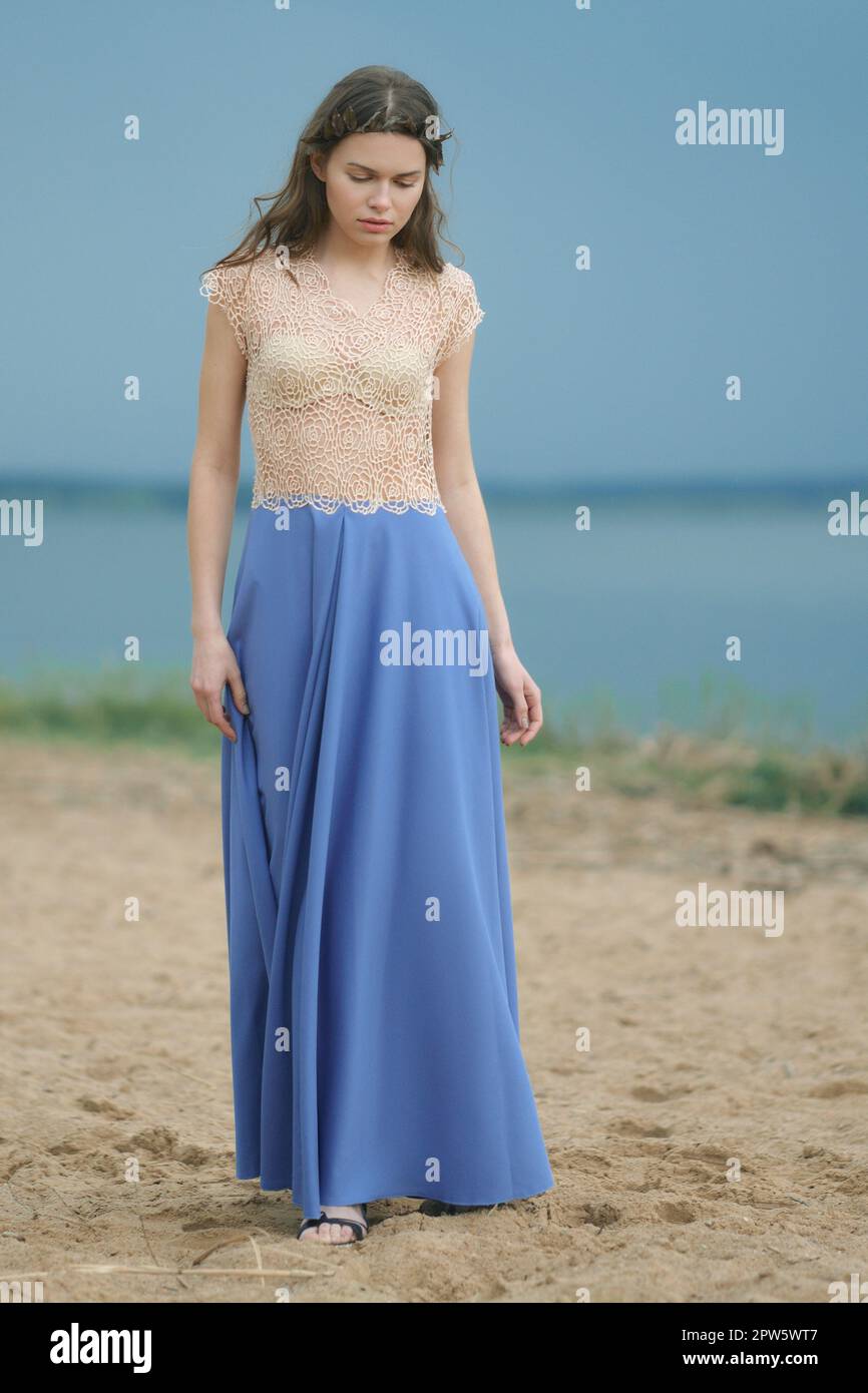 Süße junge Frau, die an einem bewölkten Tag im Sand spaziert und nach unten schaut. Sanfte Frau in langem blauem Rock und Spitzenbluse. Stockfoto