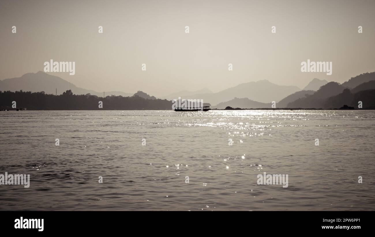 Sepiabarbene Silhouette des Slow Bootes auf dem Mekong, der an Laos und Thailand grenzt, mit Umrissen der umliegenden Berge. Stockfoto