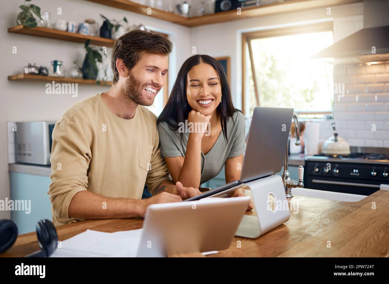 Glücklicher junger Kaukasier, der an einem Laptop arbeitet, während seine Frau neben ihm steht und sie zusammen auf den Bildschirm schauen. Ein Mann, der freiberuflich arbeitet und Showin Stockfoto