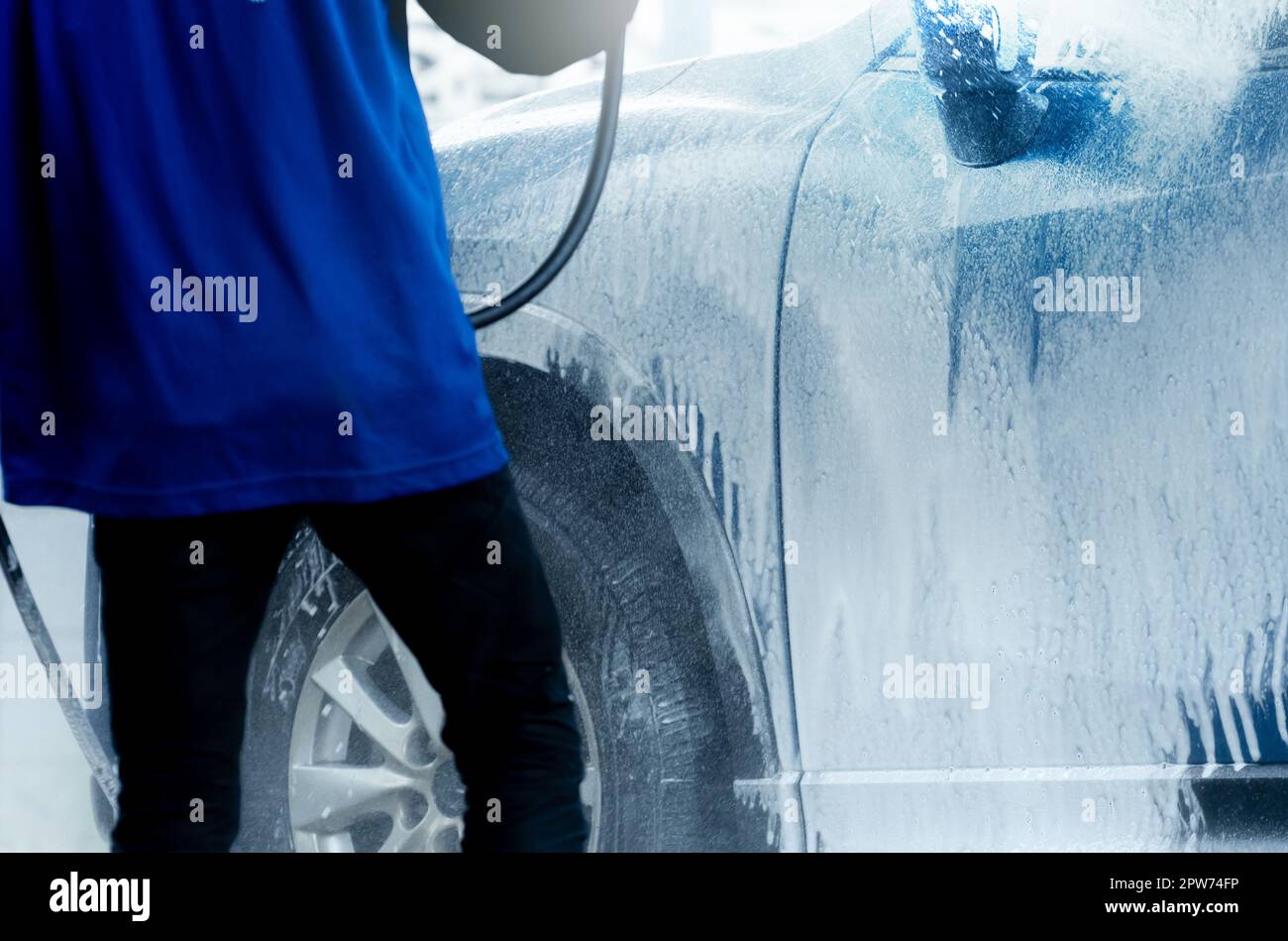 Der Arbeiter wäscht den blauen SUV mit Seifenschaum. Autowaschanlage außen. Autopflegegeschäft. Fahrzeugreinigung. Autowaschanlage-Servicekonzept. Fahrzeug mit weißem foa bedeckt Stockfoto