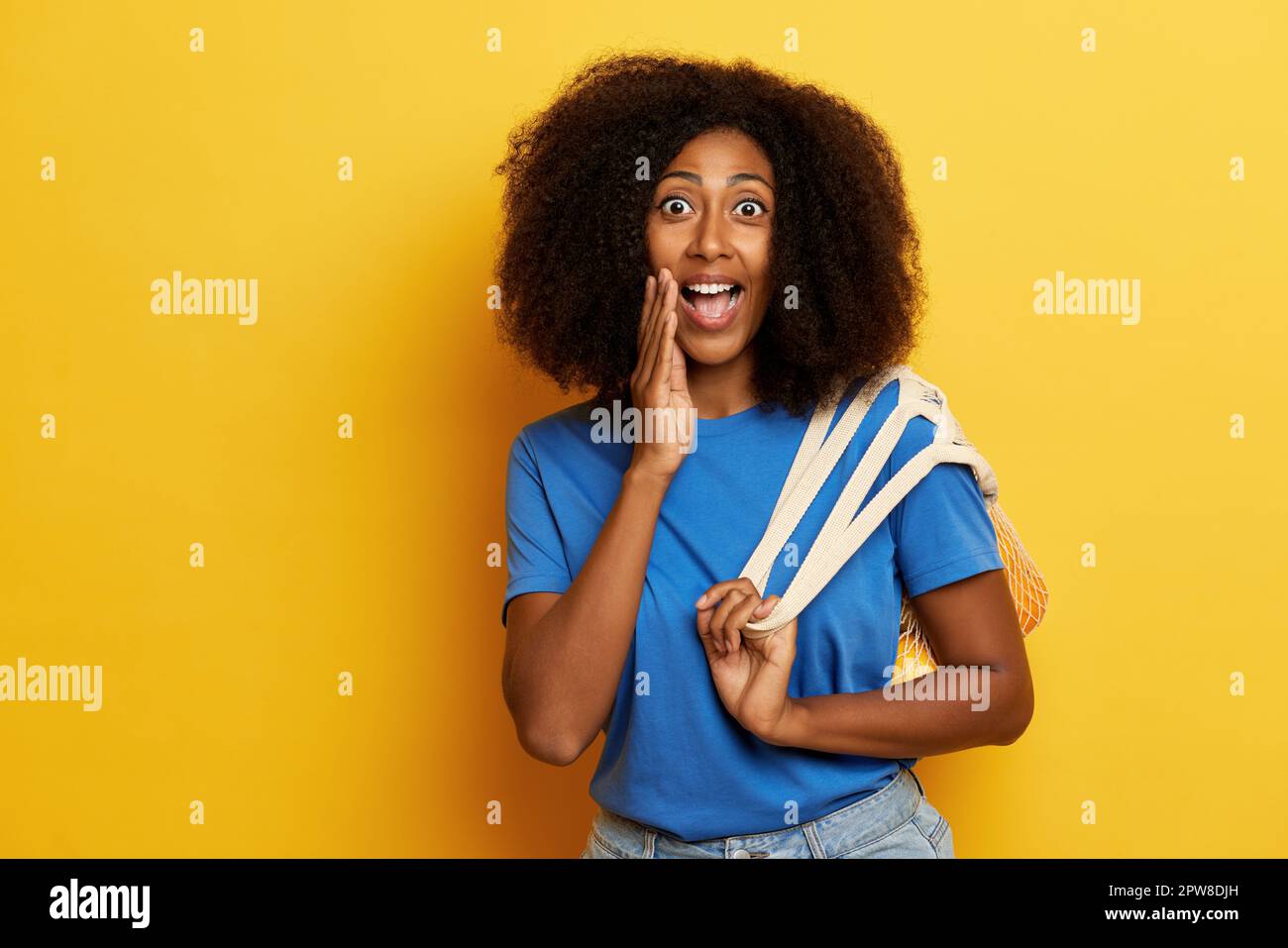 Dunkelhäutige, lockige Frau, steht neben der gelben Wand mit weit geöffneten Augen. Stockfoto