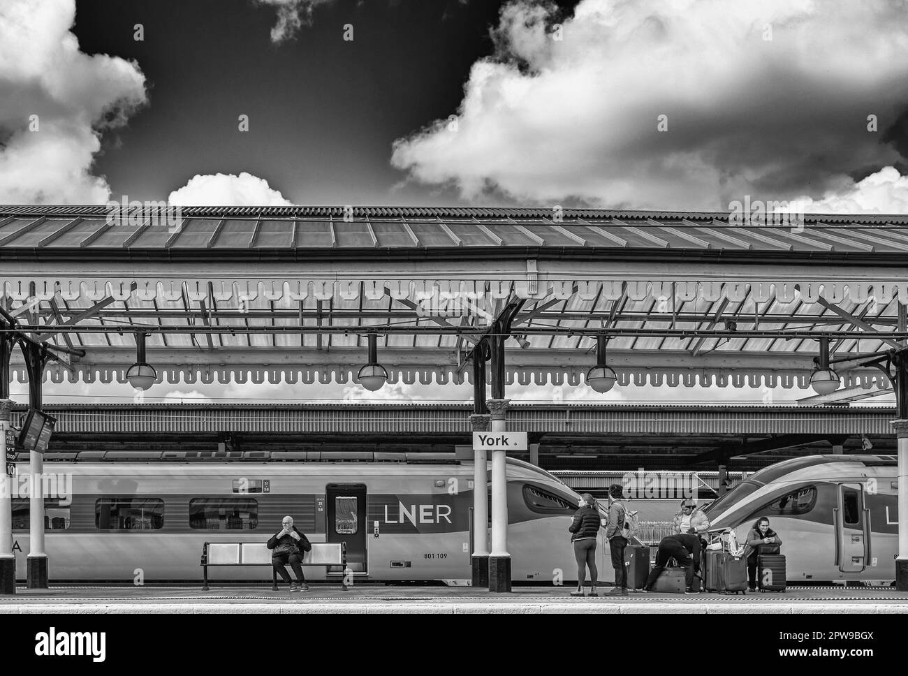 Die Passagiere warten auf einem Bahnsteig auf einen Zug. Zwei Lokomotiven befinden sich hinter ihnen und ein historisches Vordach ist über ihnen. Stockfoto