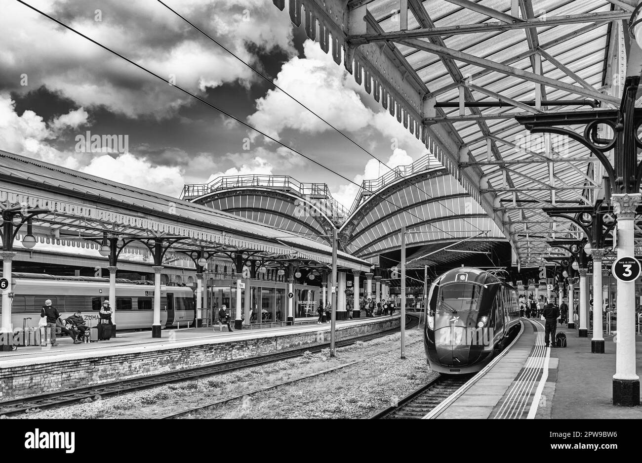 Ein Zug steht neben einem Bahnsteig mit Architektur aus dem 19. Jahrhundert. Passagiere warten auf Züge und ein Himmel mit Wolken ist oben. Stockfoto