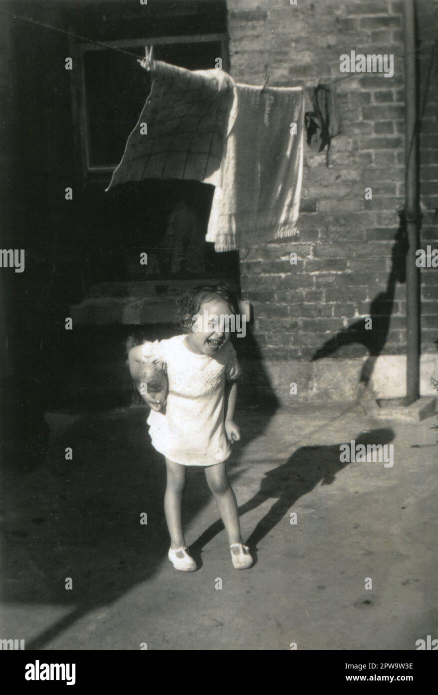 England.1930er. Ein bezauberndes Foto, das ein Kleinkind darstellt. Das junge Mädchen, das ein weißes Kleid und Sandalen trägt, steht in einem Hinterhof, klammert sich ein weiches Spielzeug unter den Arm und lacht. Handtücher und Kleidung trocknen an einer Wäscheleine über ihrem Kopf. Stockfoto