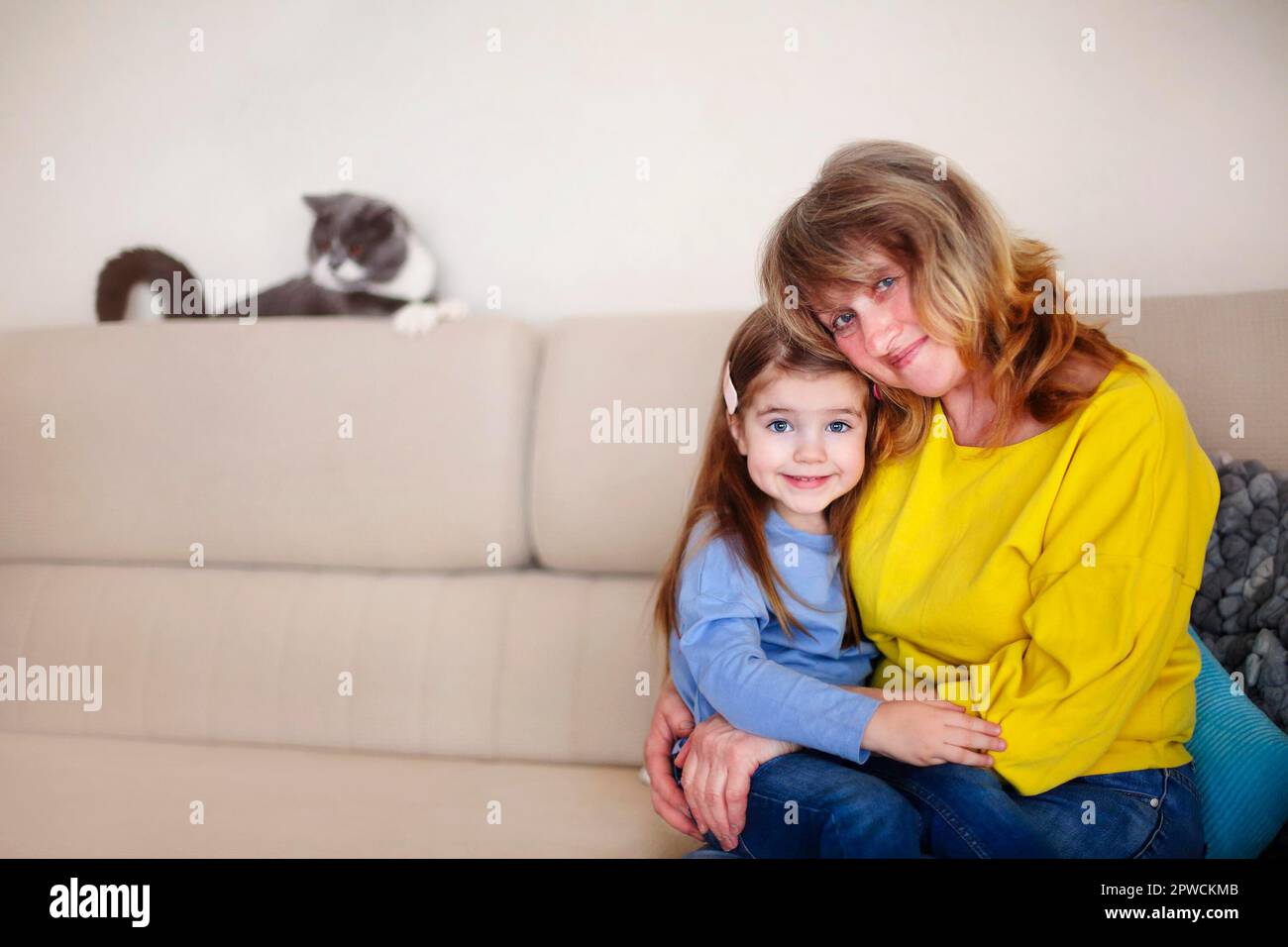 Lächelnde Großmutter mittleren Alters und kleine Enkelin, die sich umarmt und in die Kamera schaut, während sie auf dem Sofa mit einer süßen Katze im Hintergrund sitzt Stockfoto