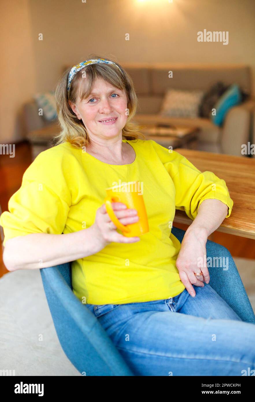Positive Frau mittleren Alters in lässigem gelben Hemd und Jeans sitzt am Tisch mit einer Tasse Tee in der Hand und schaut auf Kamera während zu Hause ausruhen Stockfoto