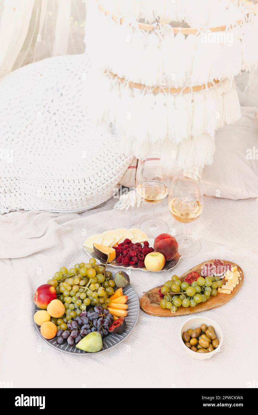 Paar Gläser mit Wein in der Nähe von Tischen mit verschiedenen platziert Frische reife Früchte auf Decke mit Kissen während des romantischen Picknicks Im Garten Stockfoto