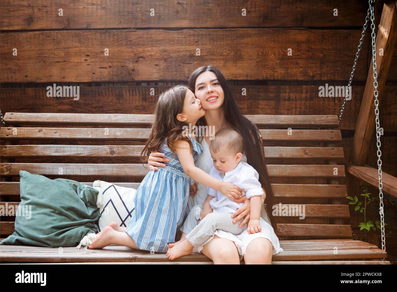 Junge, entzückte Mutter sitzt im Sessel mit entzückenden kleinen Geschwistern auf der Terrasse des Landhauses und entspannt sich am Wochenende zusammen Stockfoto