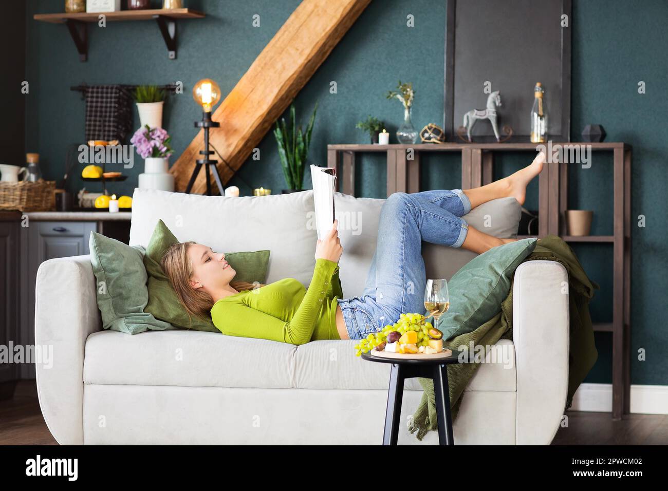 Seitenansicht einer jungen Frau beim Lesen eines interessanten Magazins im Liegen Auf der bequemen Couch neben dem Tisch mit Obst und Wein Wochenendtag zu Hause Stockfoto