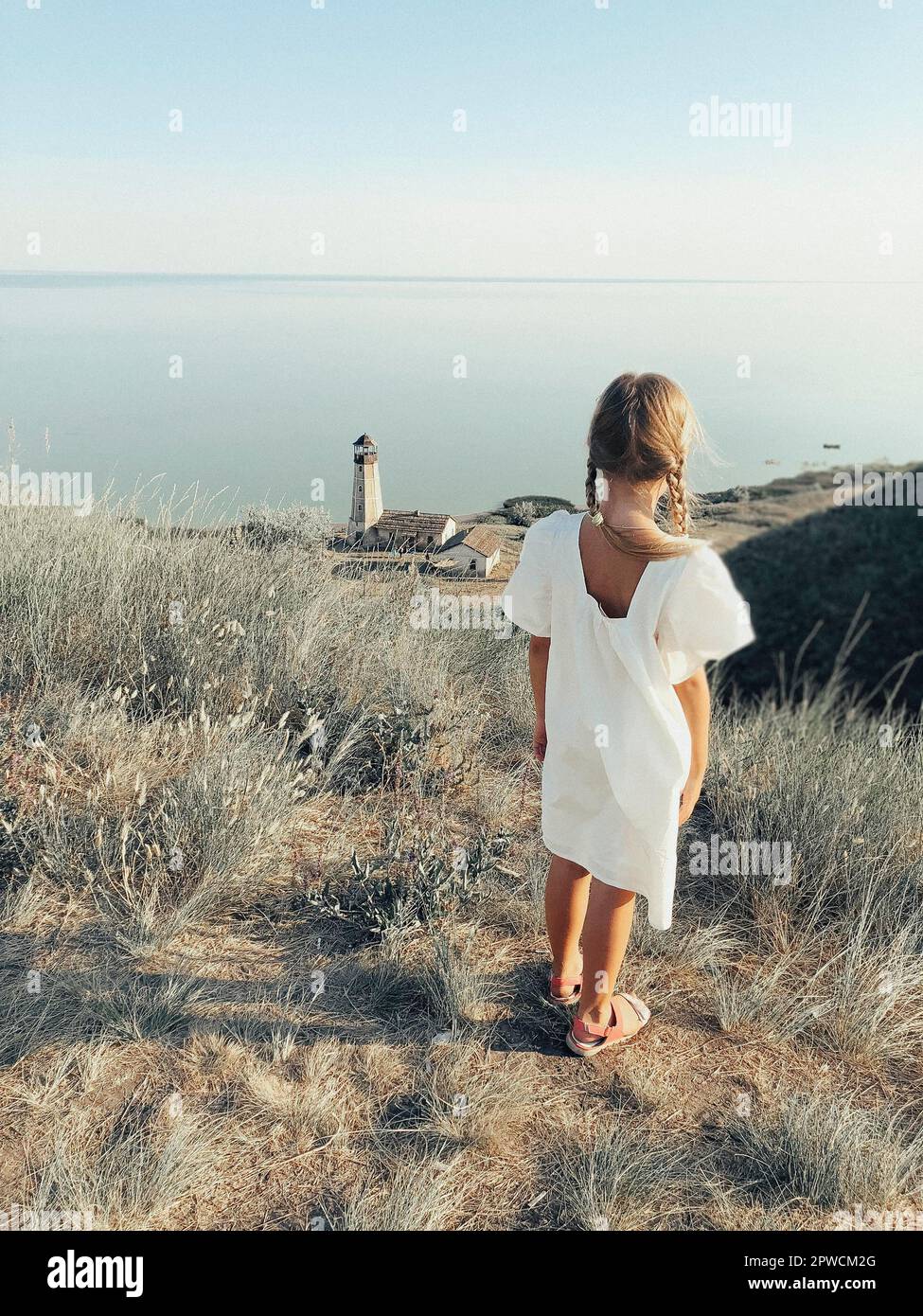 Ganzkörper-Rückansicht von einsamen unkenntlich Mädchen in weiß Sommerkleid steht auf grasbewachsenen Hügel an der Küste mit Leuchtturm Turm Stockfoto