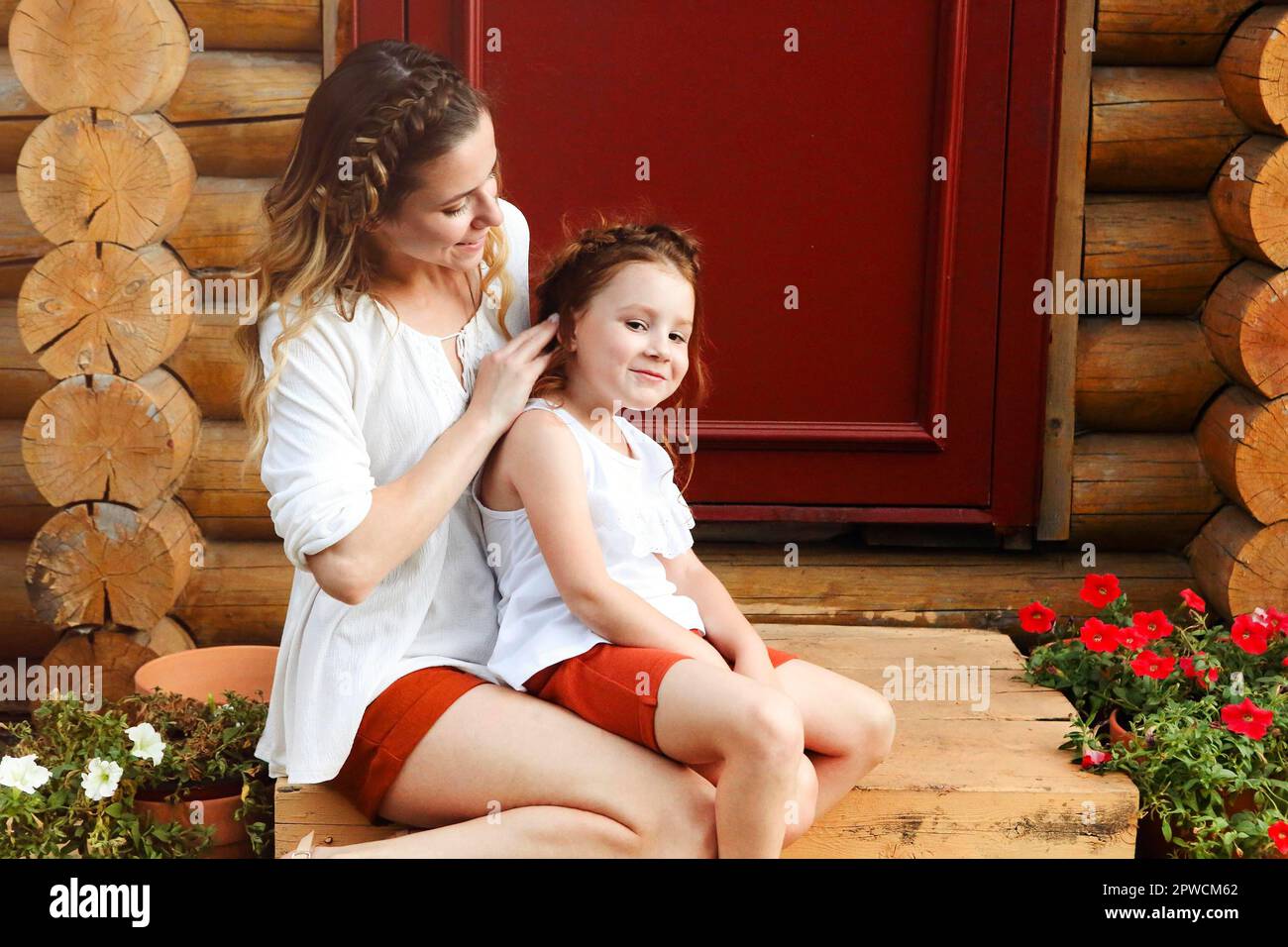 Nette kleine Rotschopf Mädchen und glücklich junge Mutter mit ähnlichen Geflochtene Frisur umarmt sich zusammen, während sie gegen die rote Tür steht Stockfoto