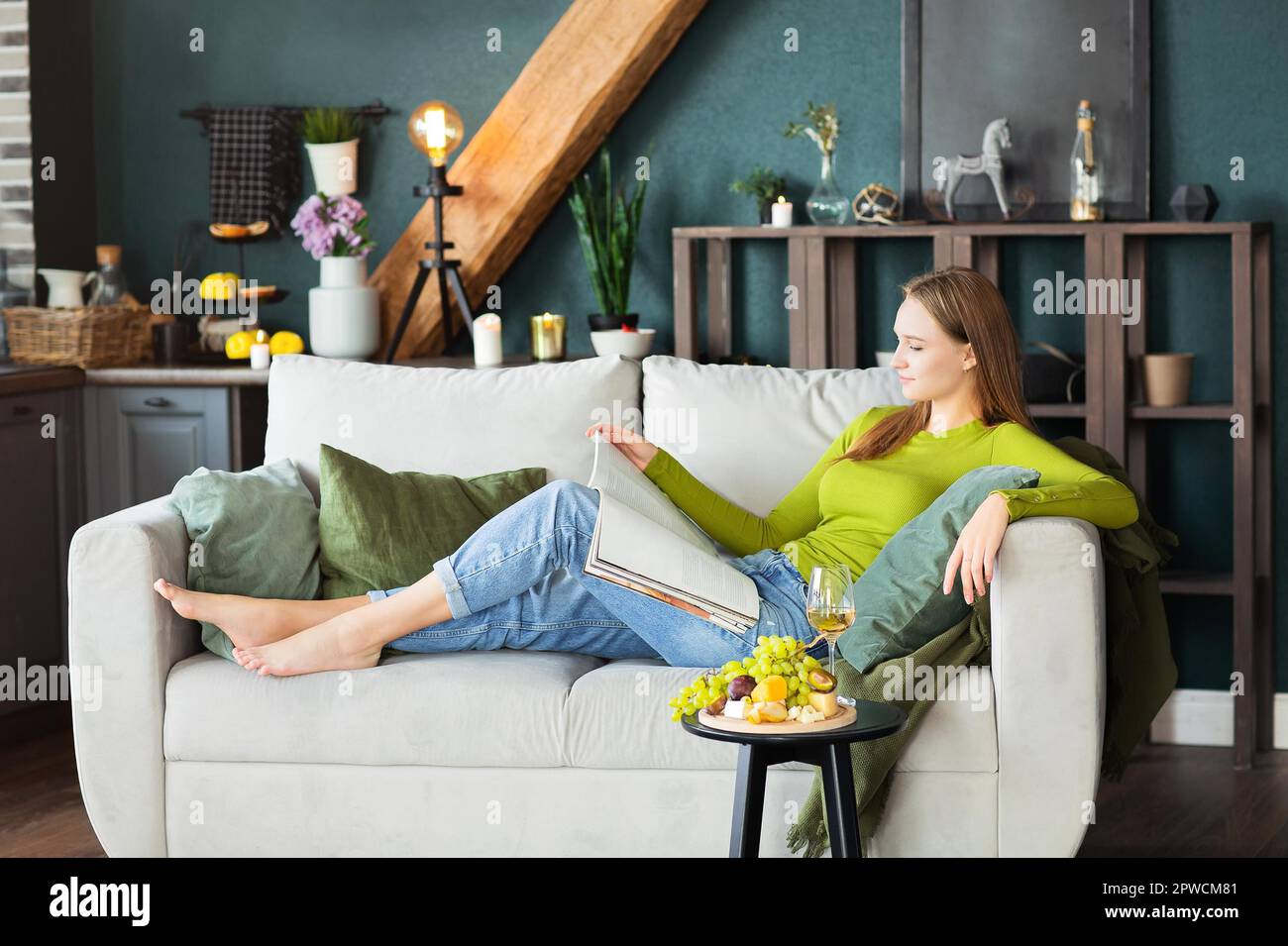Seitenansicht einer jungen Frau beim Lesen eines interessanten Magazins im Liegen Auf der bequemen Couch neben dem Tisch mit Obst und Wein Wochenendtag zu Hause Stockfoto