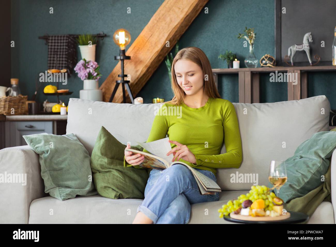 Junge blonde Frau liest interessante Zeitschrift, während sie bequem auf sitzt Couch am Tisch mit Obst und Wein am Wochenende Zu Hause Stockfoto