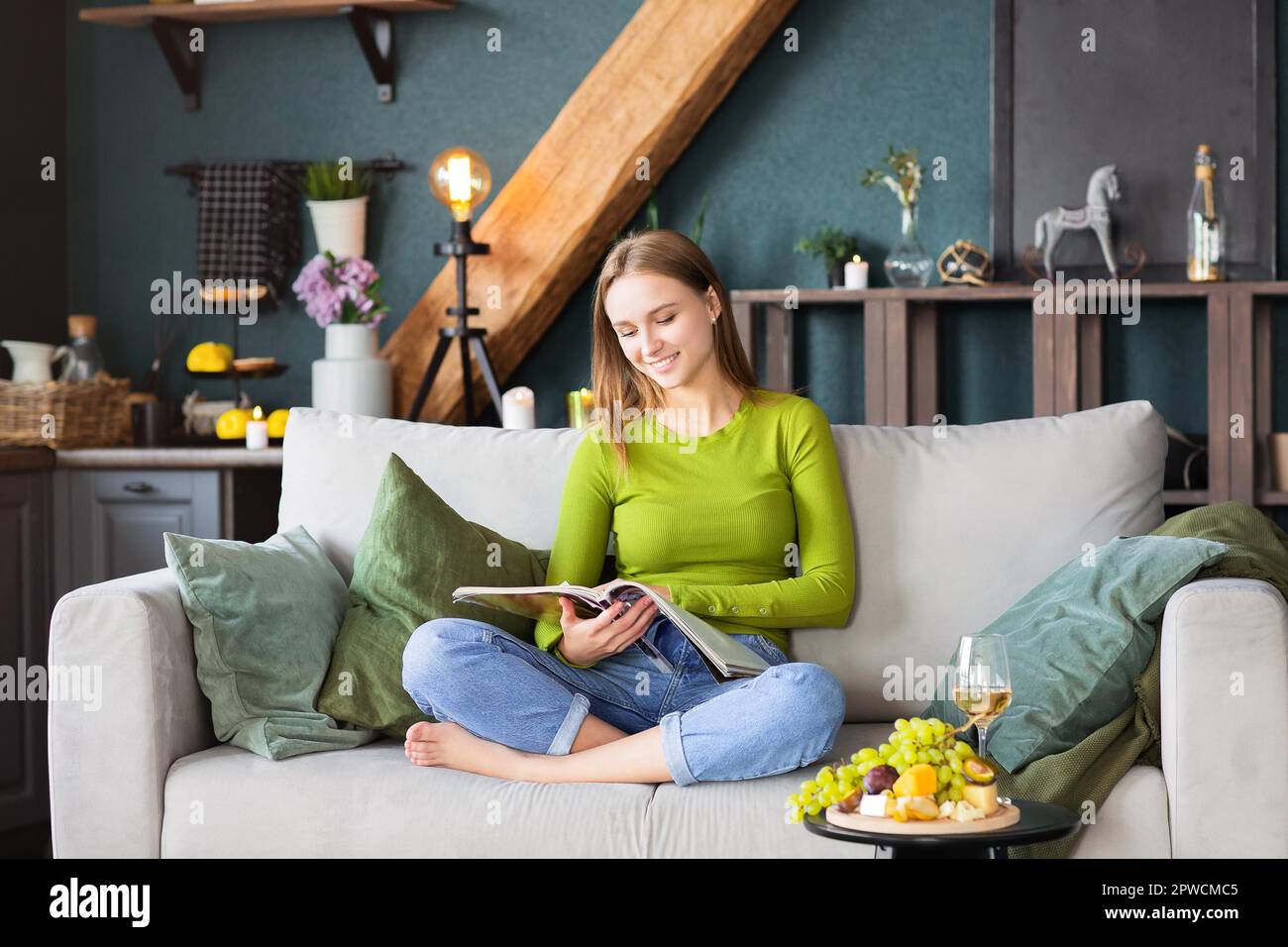 Junge blonde Frau liest interessante Zeitschrift, während sie bequem auf sitzt Couch am Tisch mit Obst und Wein am Wochenende Zu Hause Stockfoto