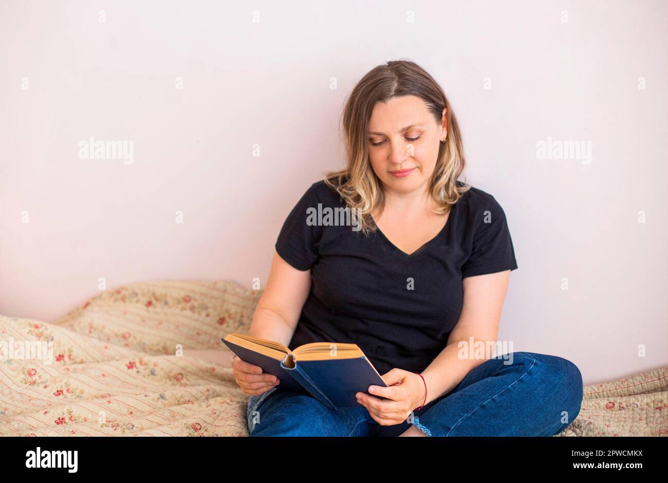 Entzückende Frau mittleren Alters in häuslicher Kleidung auf einem gemütlichen Bett im Schlafzimmer mit einer Tasse Kaffee und einem interessanten Buch liegend, während sie die Kamera betrat Stockfoto