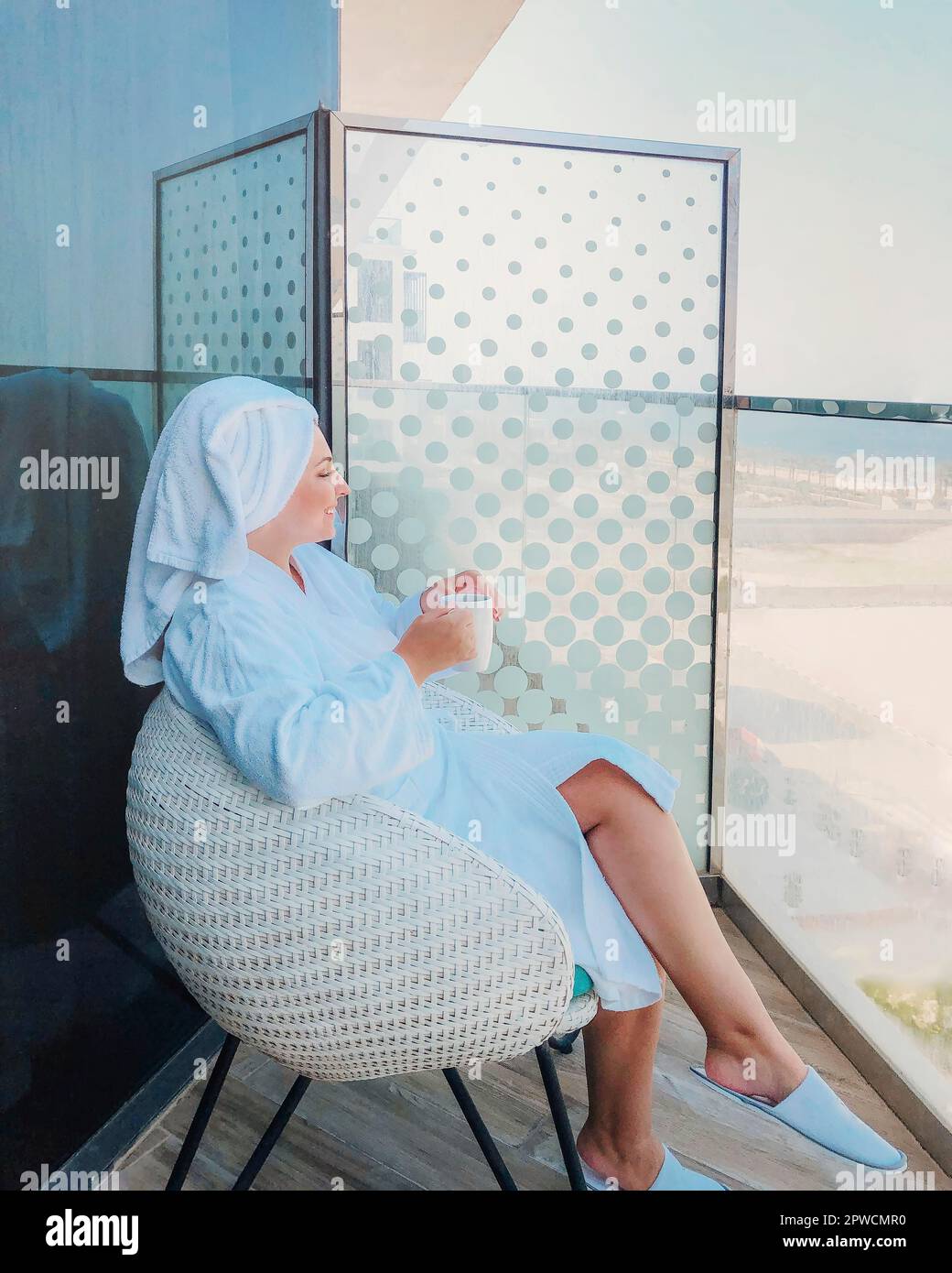 Seitenansicht einer jungen Frau im weißen Bademantel und einem Handtuch auf dem Kopf, das auf einem Sessel sitzt und eine Tasse Kaffee auf dem Balkon Stockfoto