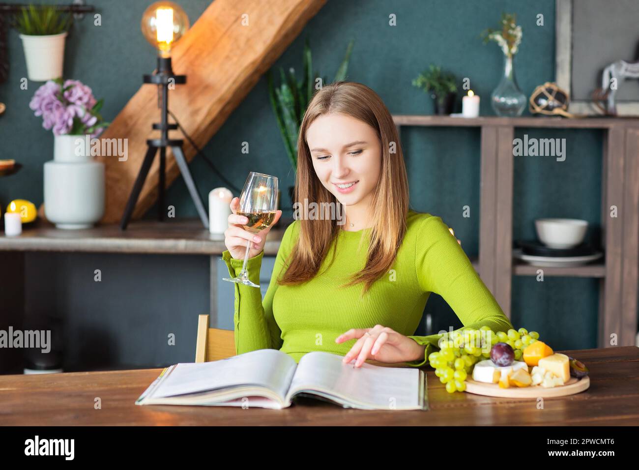 Junge Frau liest am Holztisch ein Interessantes Magazin mit Obst und Wein an einem Wochenende zu Hause Stockfoto