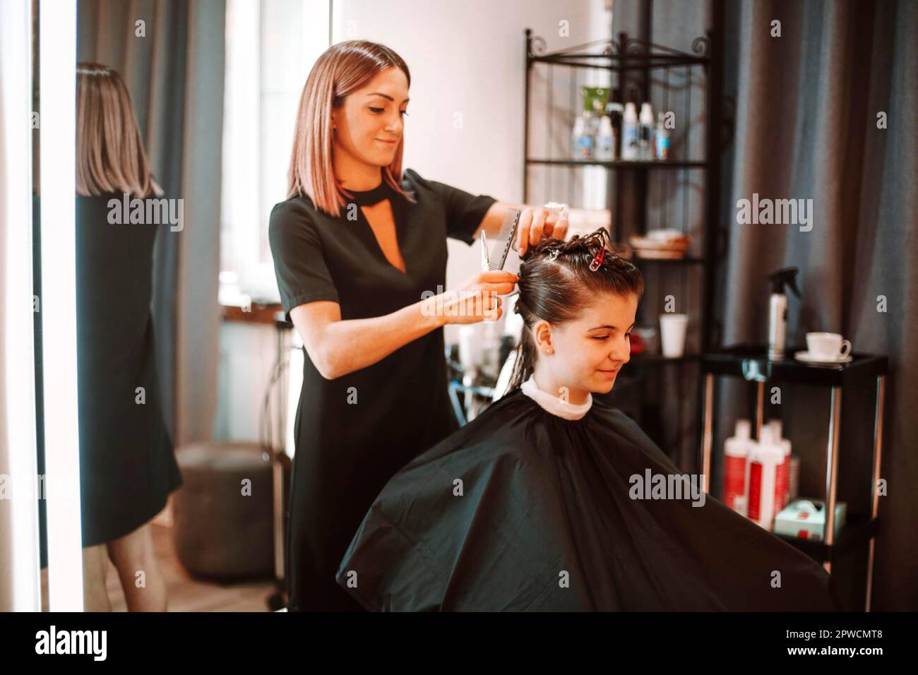 Positive Friseurin bei der Arbeit im Schönheitssalon, professioneller, lächelnder Friseur, der eine Klientin für den Haarschnitt vorbereitet, Haare anheften Stockfoto