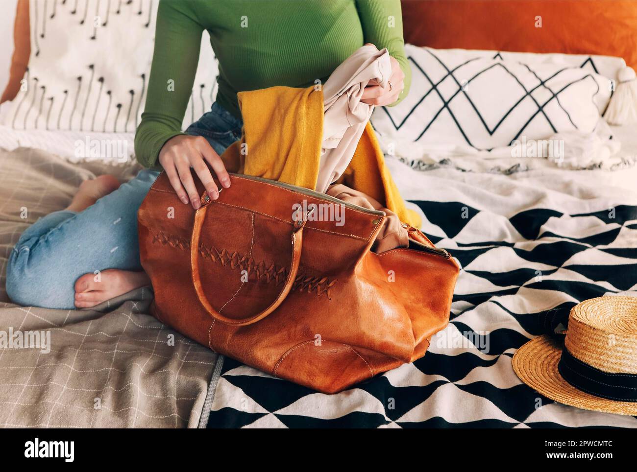 Junge schöne Frau in lässiger Kleidung, die zu Hause auf dem Bett sitzt und ihren gelben Herbstschal in eine Lederhandtasche steckt, um draußen spazieren zu gehen Stockfoto