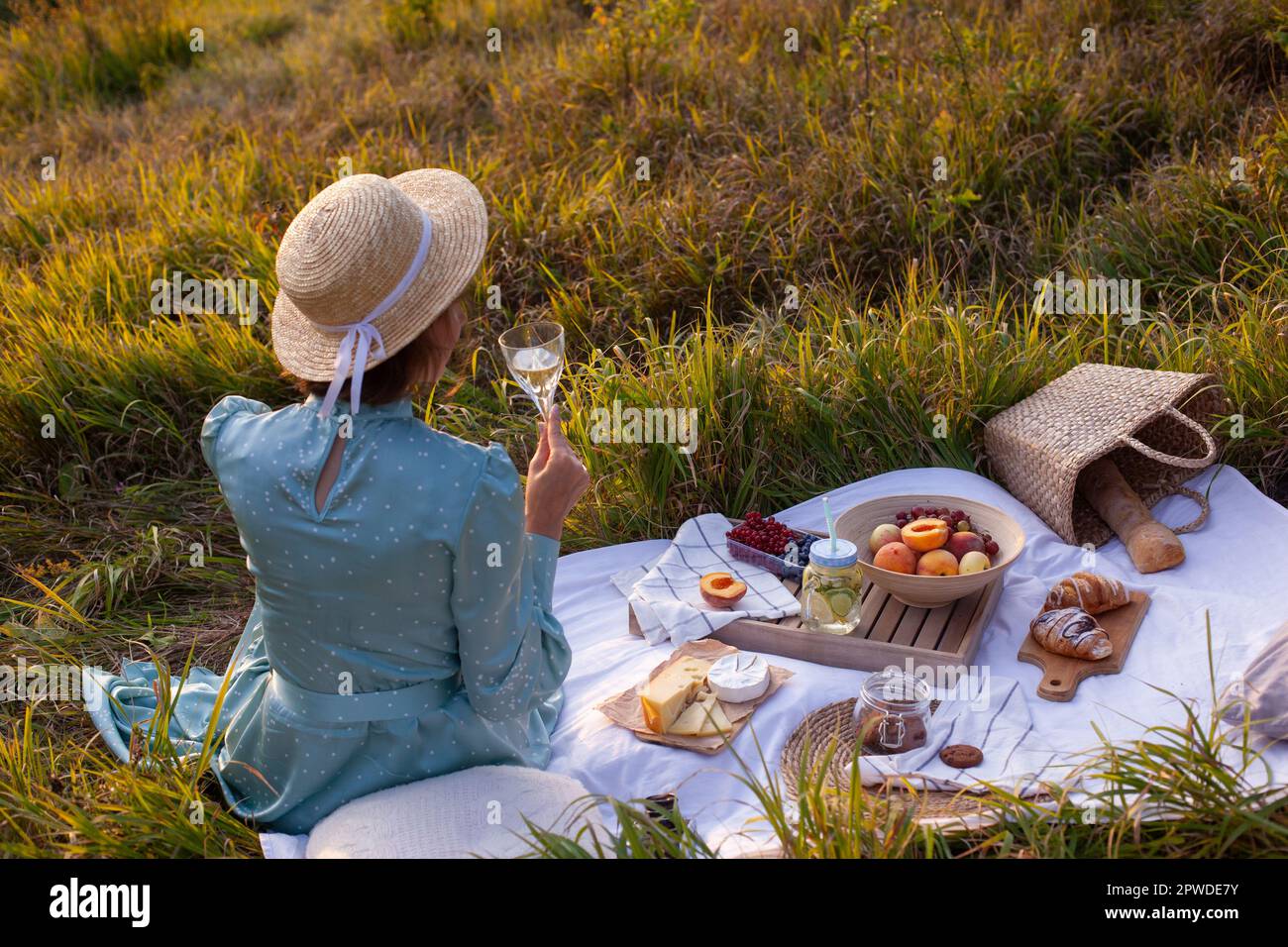 Eine Frau in blauem Kleid und Strohhut mit kurzen Haaren, die auf einer weißen Decke sitzt, ein Glas hält und Weißwein trinkt. Das Konzept, ein Picknick in einem zu machen Stockfoto