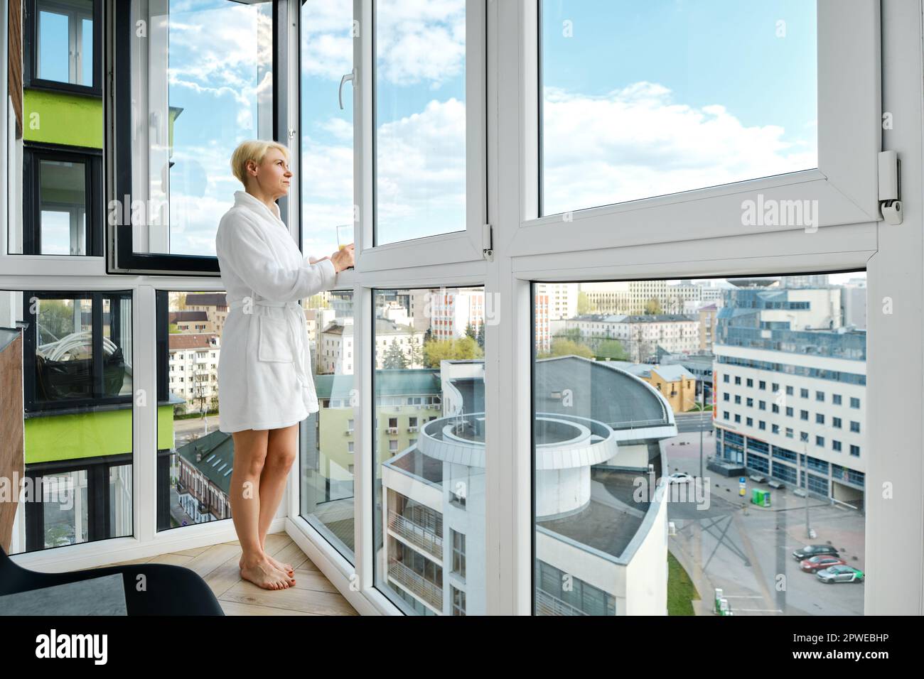 Eine reife, sorgenfreie Frau steht barfuß in einem Bademantel auf ihrem Balkon und genießt einen Orangensaft. Allein in ihrem neuen Hochhaus und mit Blick auf das Haus Stockfoto