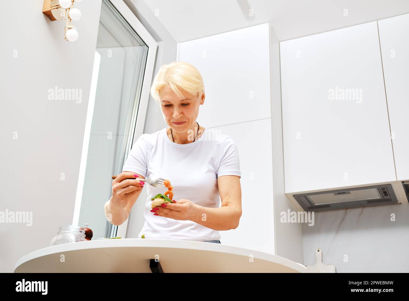 Blick aus dem niedrigen Winkel auf eine Frau mittleren Alters, die während des Frühstücks salzigen Lachs auf ein Sandwich legte Stockfoto