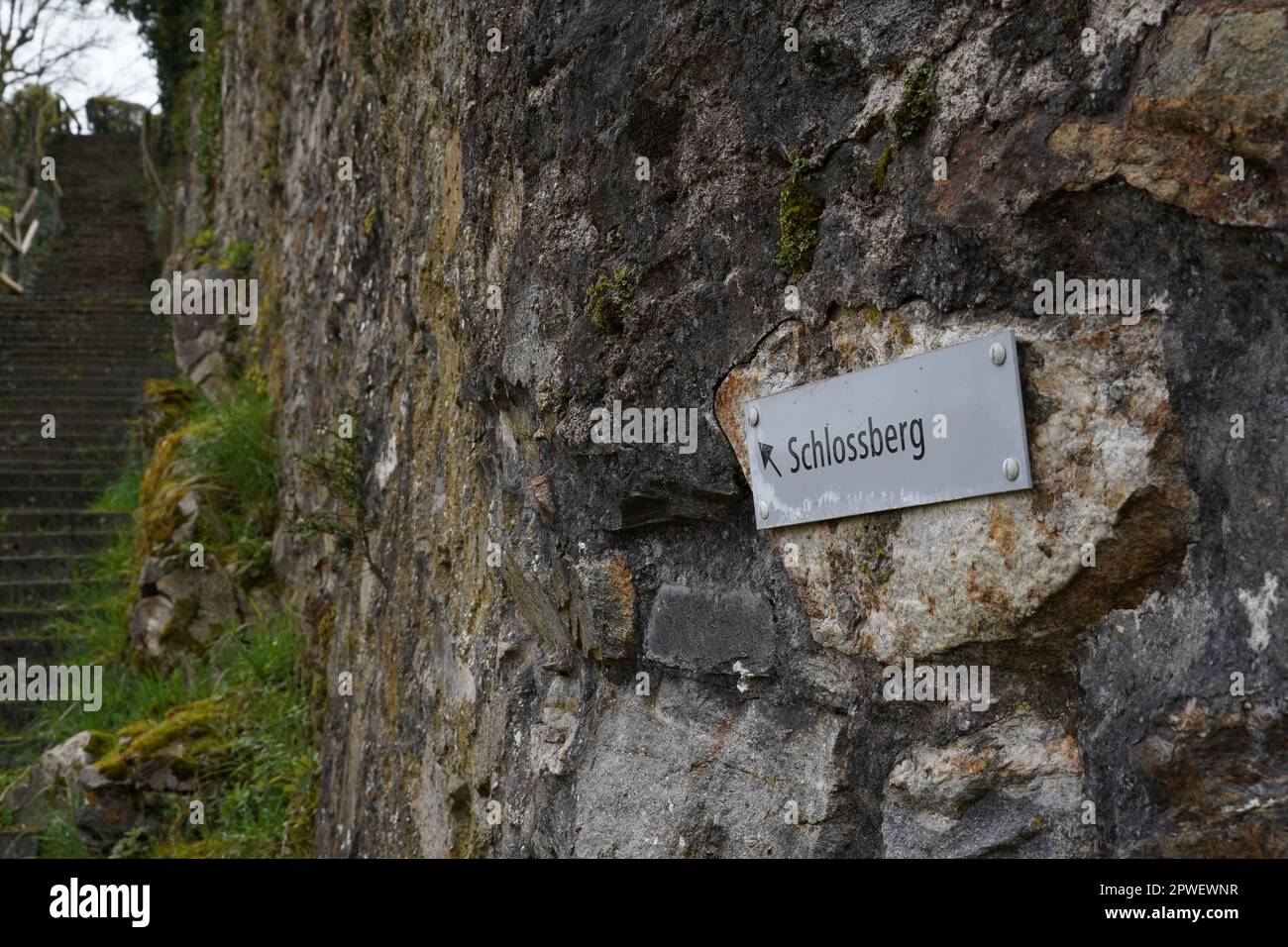 Steinmauer mit Metallschild mit Pfeil und deutscher Inschrift Schlossberg mit Bedeutung Burggestein. Stockfoto