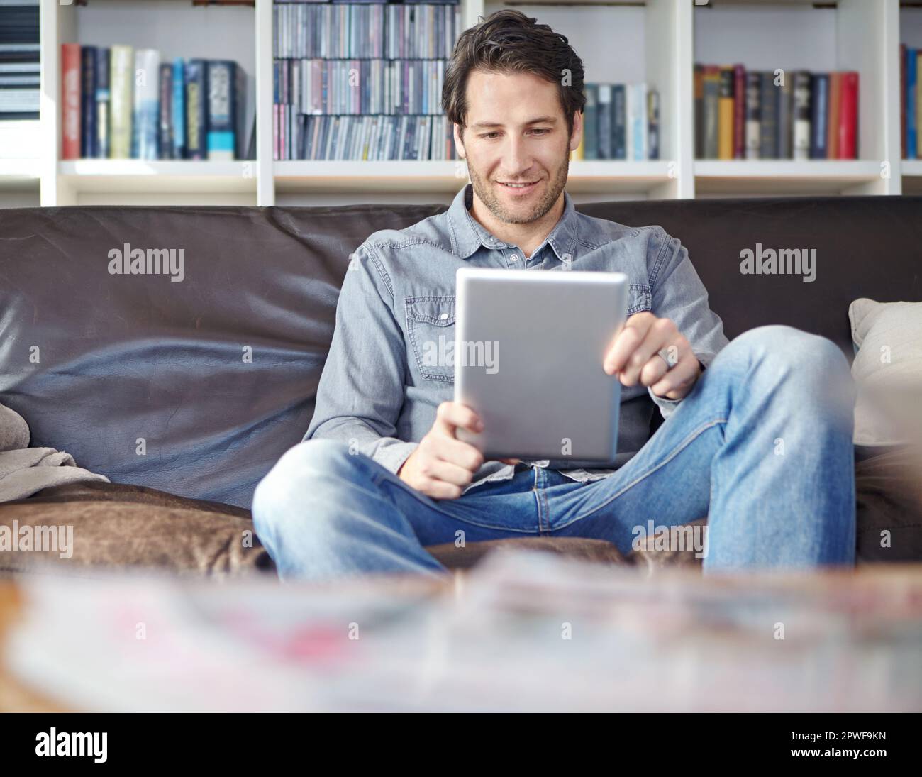 Er hat sich am Wochenende unterhalten. Ein gutaussehender junger Mann, der auf einer Couch sitzt, während er ein digitales Tablet benutzt. Stockfoto