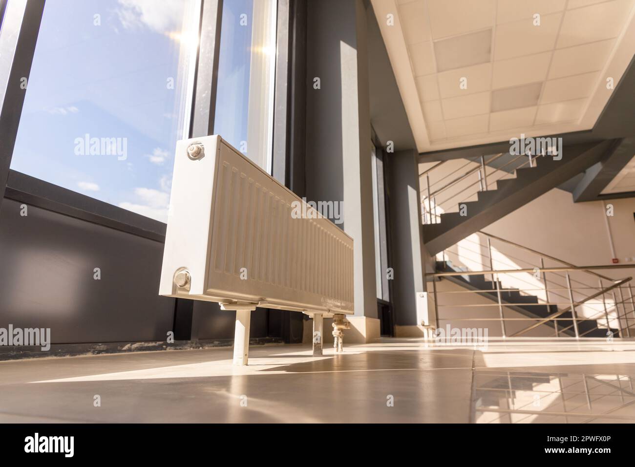 Vertikal installierter Zentralheizungskühler auf dem Boden an der vollständig verglasten Außenwand des Bürogebäudes. Ein weißer Heizkörper in einem Raum mit einem Stockfoto