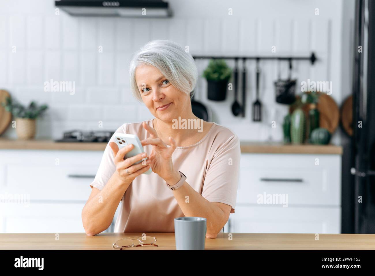 Eine wunderschöne, positive, grauhaarige Frau mittleren Alters, sitzt zu Hause in der Küche, nutzt ihr Smartphone, um in sozialen Netzwerken zu kommunizieren, Nachrichten zu lesen, macht eine Online-Bestellung, lächelt vor der Kamera Stockfoto