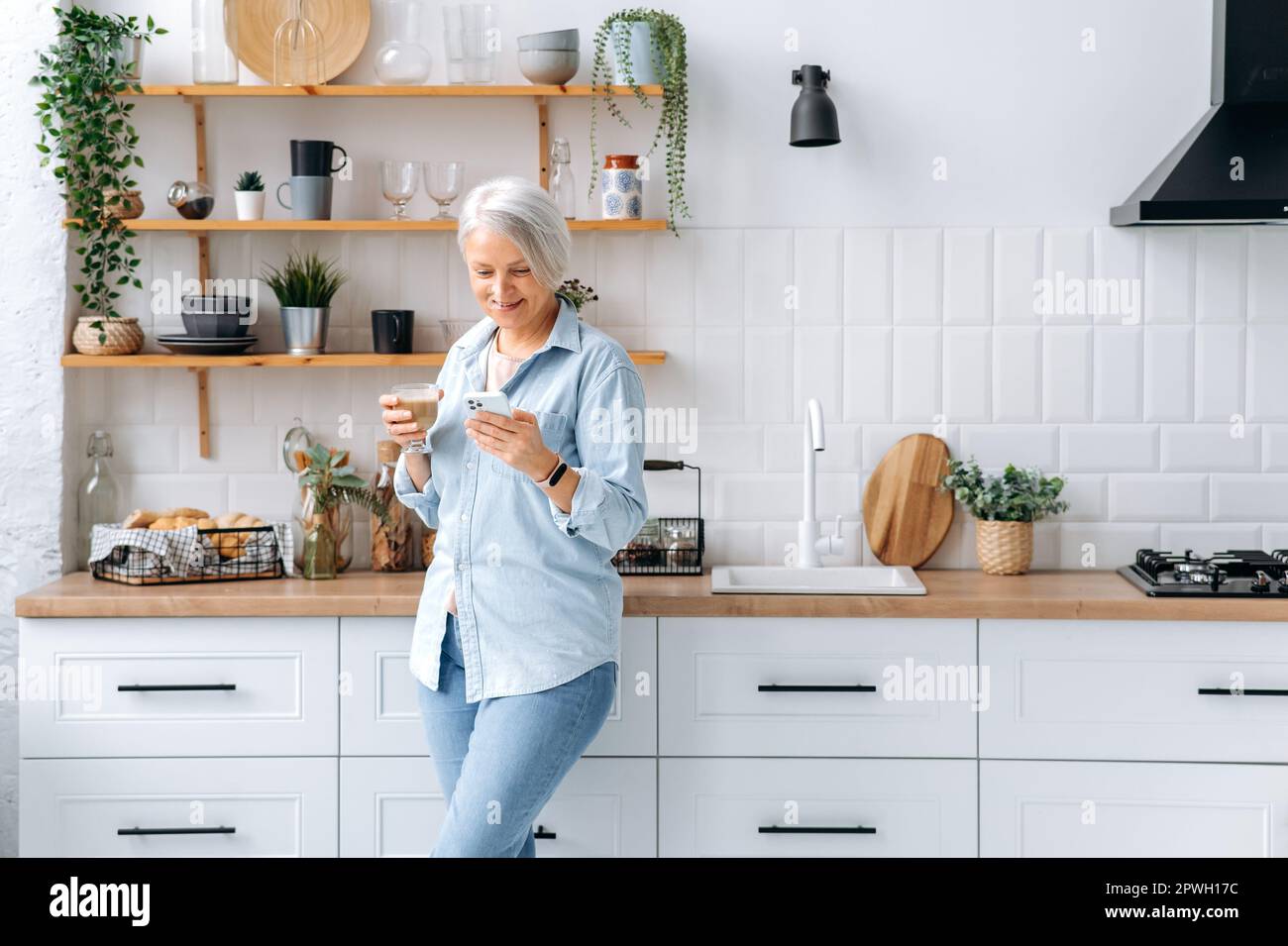 Wunderschöne, fröhliche, grauhaarige Frau mittleren Alters, steht zu Hause in der Küche, hält Smartphone und ein Glas Kaffee, schickt Nachrichten in sozialen Netzwerken, liest Nachrichten, macht eine Online-Bestellung, lächelt Stockfoto