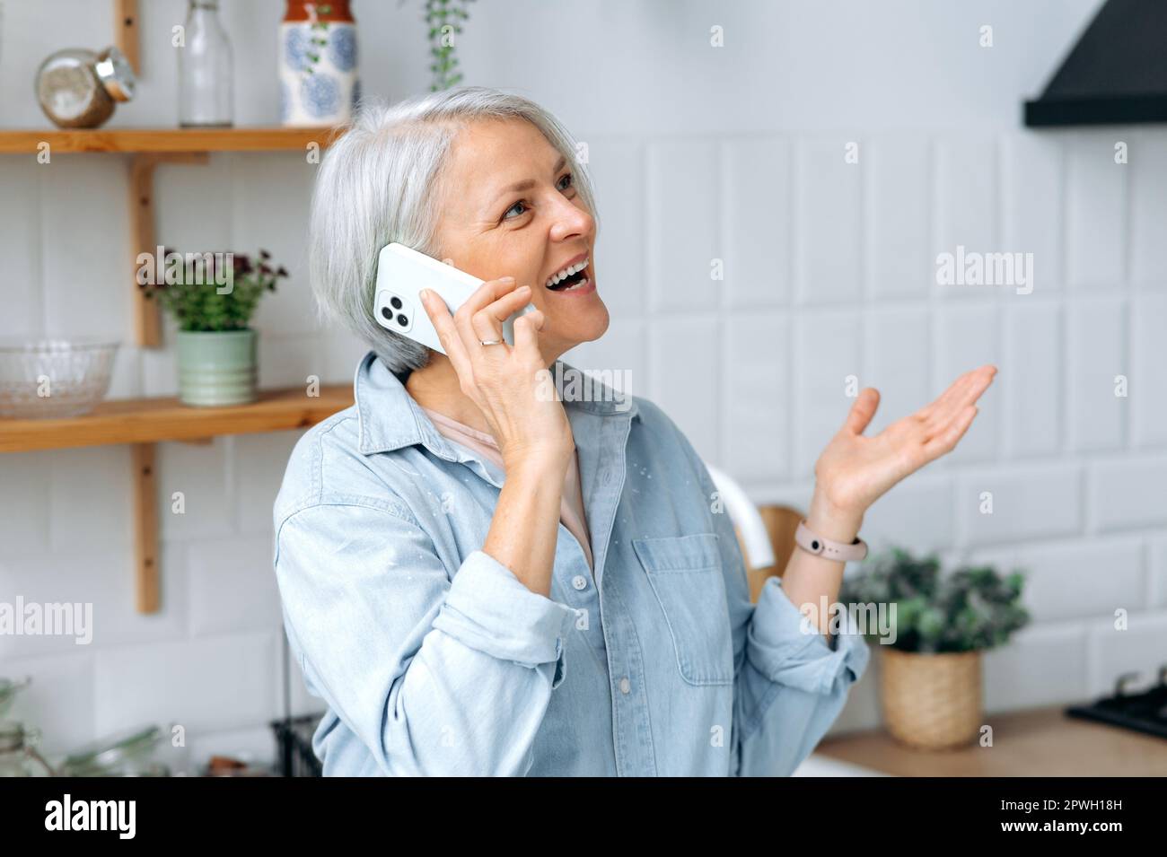 Ein Anruf. Positive weiße, reife, grauhaarige Frau, zu Hause in der Küche stehen, mit einem Smartphone mit einem Freund oder der Familie sprechen, nette Arbeitsgespräche führen, wegschauen, mit der Hand Gesten machen, lächeln Stockfoto