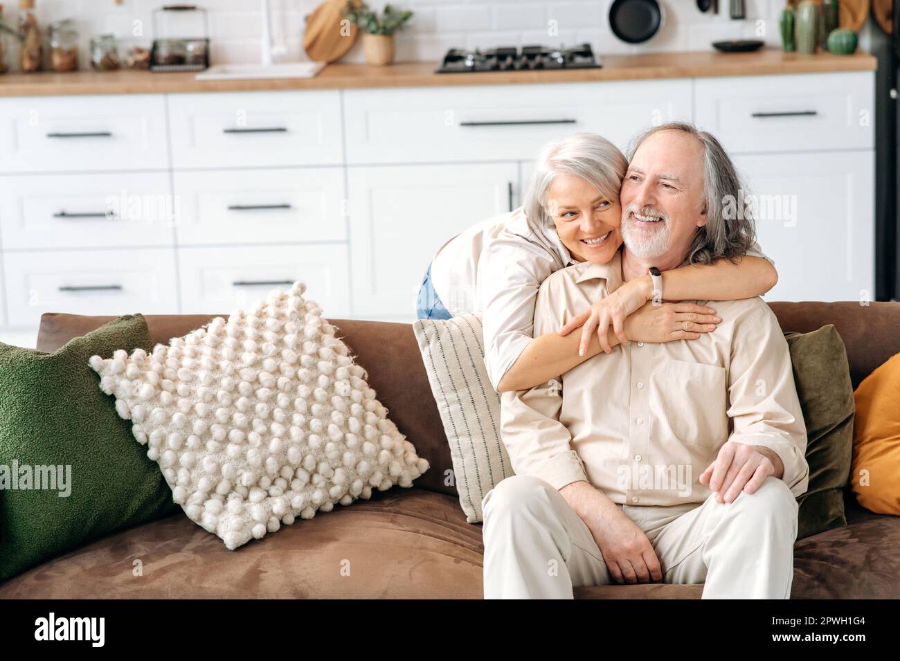 Foto von glücklichen weißhäutigen grauhaarigen Ehepartnern im Rentenalter, Ehemann und Ehefrau verbringen Zeit zusammen im Wohnzimmer, eine Frau umarmt ihren geliebten Mann, sitzt auf dem Sofa, sie lächeln, freuen sich, zusammen zu sein Stockfoto