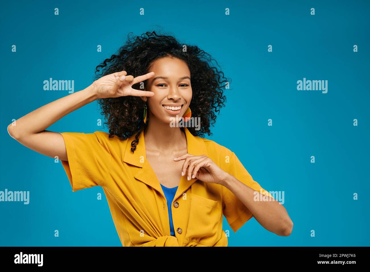 Das fröhliche, schwärzende Mädchen freut sich und hat Spaß in hellen Sommerkleidung, die mit ihren Fingern ein V-Schild auf blauem Hintergrund zeigt Stockfoto