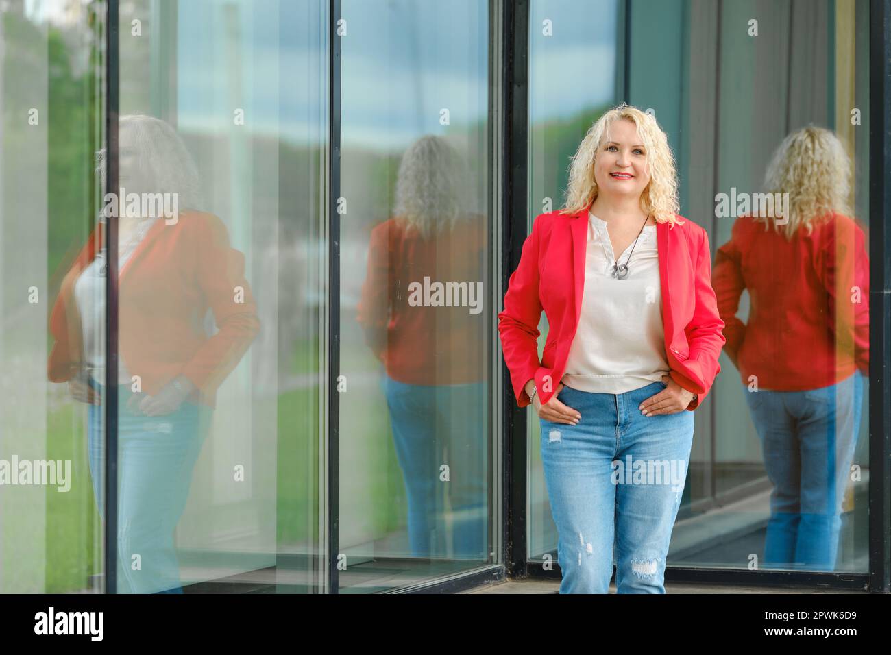 Süße Seniorin in roter Jacke, Bluse und Jeans, die neben der Glaswand des Gebäudes stehen Stockfoto