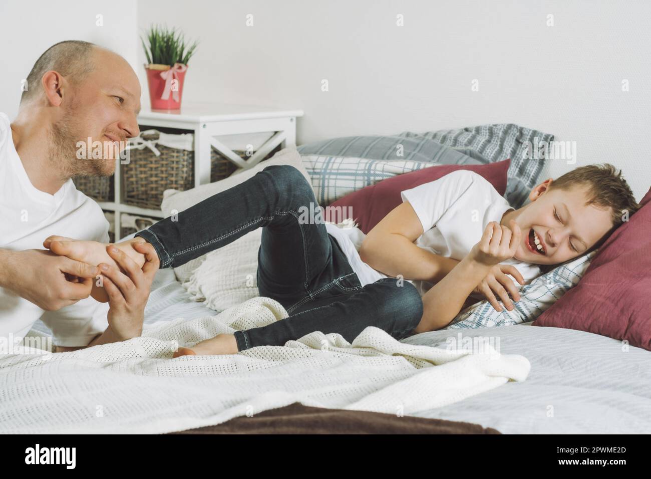 Vater spielt mit seinem Sohn 7-10 auf dem Bett. Papa kitzelt die Füße der Kinder. Familie, die Spaß hat Stockfoto