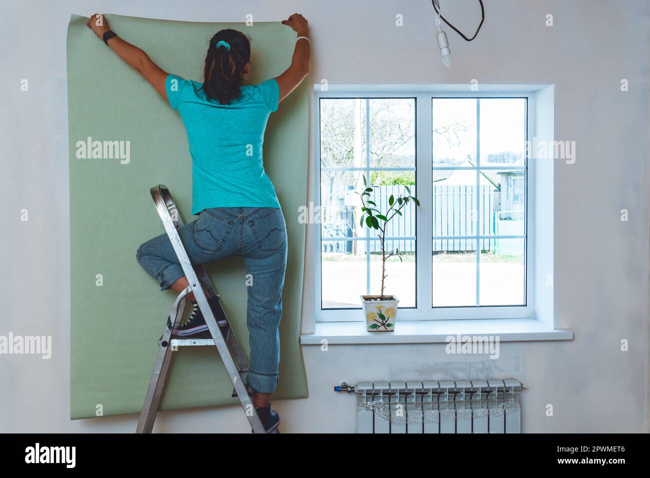 Frau in Jeans und türkisfarbenem T-Shirt, die auf einer Stehleiter steht und ein Stück Tapete hält. Renovierung und Verbesserung des Hauses. Stockfoto