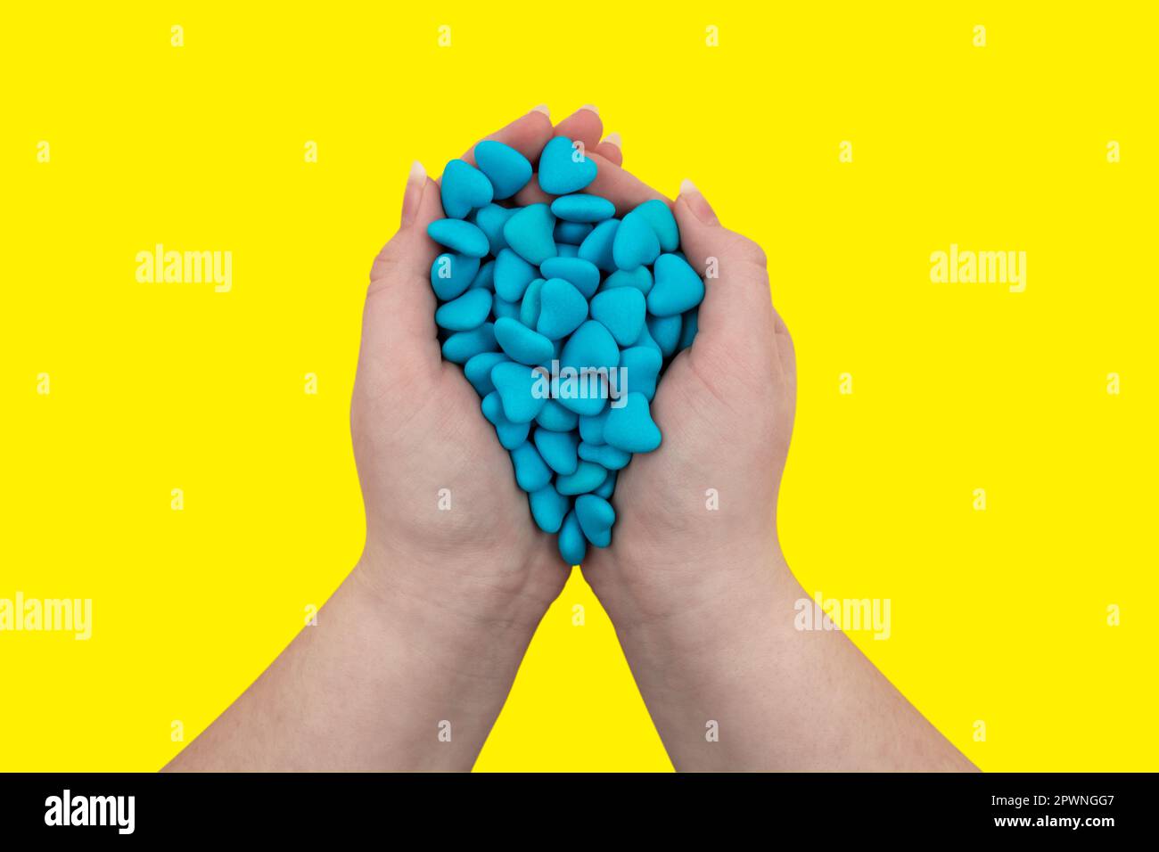 Eine Handvoll blauer herzförmiger Süßigkeiten in Händen isoliert auf gelbem Hintergrund. Staatssymbole der Ukraine. Nahaufnahme, Draufsicht Stockfoto