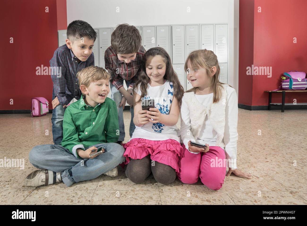 Schulkinder sitzen auf dem Boden und schauen auf Handys im Flur neben Schließfächern, Bayern, Deutschland Stockfoto