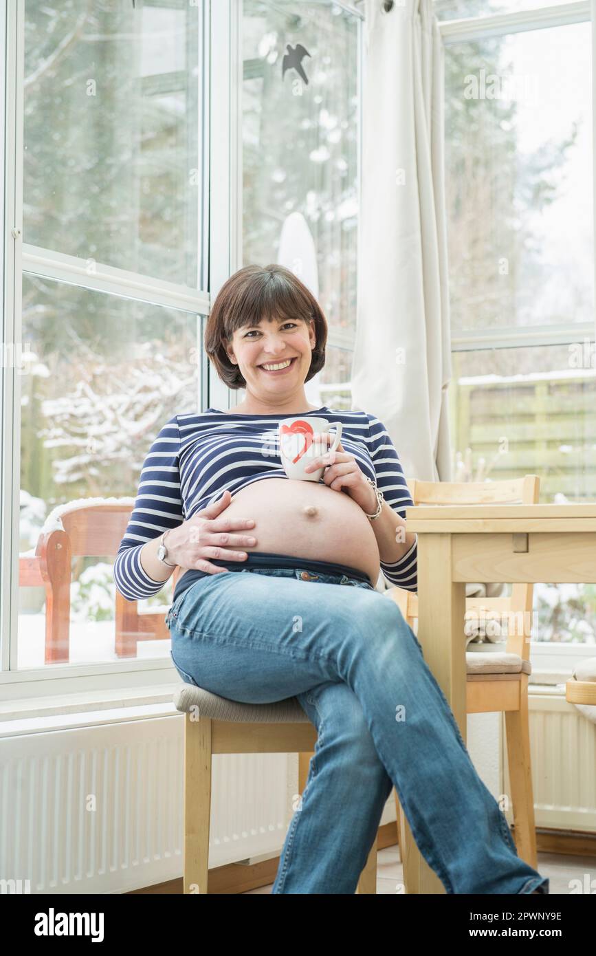 Porträt einer schwangeren Frau, die mit einer Kaffeetasse auf einem Stuhl sitzt Stockfoto